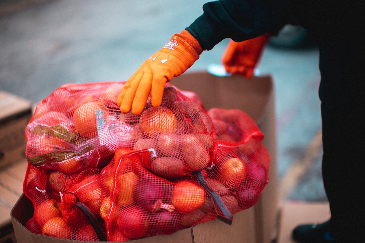 A worker wearing gloves organizes fresh apples in net bags within a cardboard box outdoors.